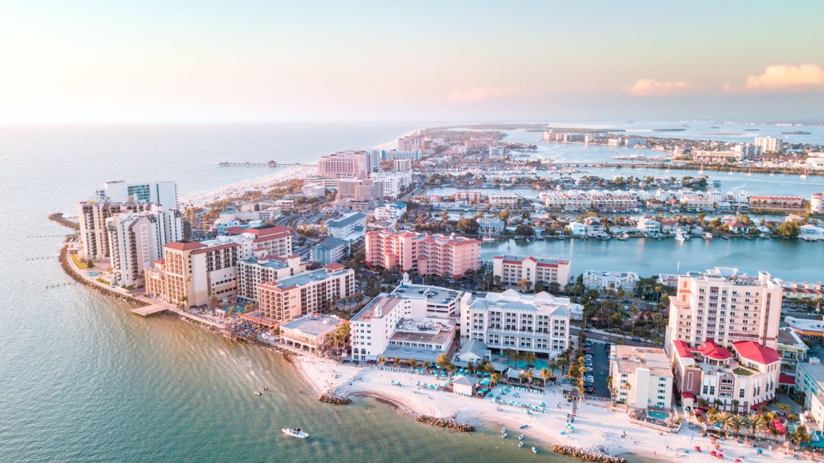 Aerial view of a seaside city with high-rise buildings, a curved shoreline, and calm water at sunset/light pastel tone.