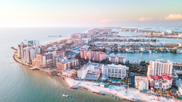 Aerial view of a seaside city with high-rise buildings, a curved shoreline, and calm water at sunset/light pastel tone.