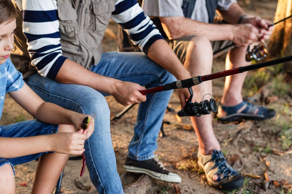 BeachesUSA-PeopleFishing-Shutterstock | Beaches USA Rentals & Management, LLC People sitting outdoors and preparing fishing gear, with a red fishing rod and reel in focus on the right side of the image.
